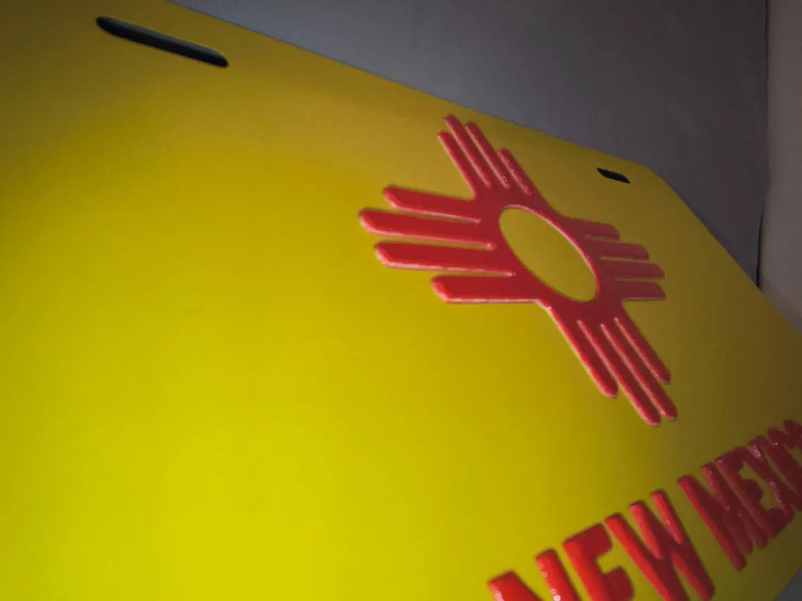 Angled close-up of a yellow aluminum vanity plate with a raised red Zia symbol and New Mexico lettering.